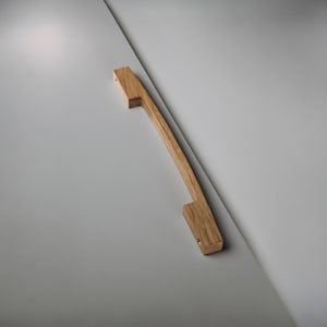 Op de afbeelding: Een gebogen houten kastgreep met een natuurlijke afwerking. De greep is gemaakt van lichtgekleurd hout en heeft aan elk uiteinde een rechthoekige vorm. De greep is gemonteerd op een lichtgrijze ondergrond.