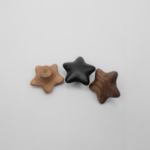 Puede incluir: Tres pomos de madera en forma de estrella en tonos marrones y negros. Uno es marrón claro con un tornillo, otro es negro y el último es marrón más oscuro con vetas de madera. Estos pomos son probablemente para muebles o decoración.