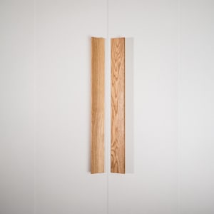 Può includere: Due maniglie verticali in legno con finitura naturale venata, montate su un'anta bianca. Le maniglie sono rettangolari e hanno una forma leggermente curva, aggiungendo un tocco di design organico all'estetica minimalista.