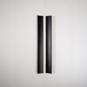 May include: Two black wooden cabinet pulls with a vertical design. The pulls are mounted on a white cabinet door. The pulls have a rectangular shape with a beveled edge.