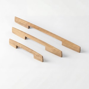 May include: Three wooden cabinet pulls in varying lengths, arranged diagonally against a white background. The pulls are a light brown color, with a rectangular shape and a handle on each end. These are ideal for kitchen or bathroom cabinets.