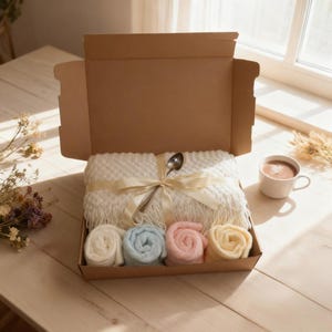 May include: A cream-coloured blanket tied with a ribbon and a spoon in an open gift box. Below are four rolled blankets in pastel shades: white, blue, pink, and yellow. A mug of coffee sits nearby. The box is open, revealing the contents.