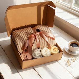 May include: A cardboard gift box containing a beige knitted blanket tied with a peach ribbon. Rolled skeins of yarn in pink, grey, and yellow are also in the box. A cup of coffee on a saucer is placed nearby.