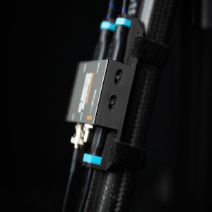 以下が含まれることがあります： 複数のケーブルとコネクタを備えた黒い金属製デバイスのクローズアップ。デバイスは、ベルクロストラップ付きのテクスチャードブラックケーブルに取り付けられています。ケーブルとデバイスには青色のアクセントが見えます。