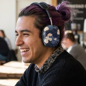May include: Black and silver headphones with earcups featuring a blue and white wave and cloud pattern. The headphones are worn by a person with purple hair. The person is smiling and wearing a black sweater.