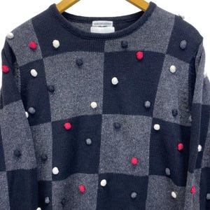 May include: A crew neck sweater with a checkerboard pattern in shades of gray and navy blue. The sweater is adorned with small, round pom-poms in red, white, and gray. The label reads "NAME THE DESIGNER".