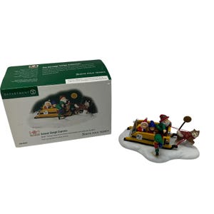 Puede incluir: Una figura de Department 56 School Sleigh Express de la colección Heritage Village. El trineo amarillo transporta a tres niños y a Papá Noel, tirados por un perro husky. Un elfo verde está en la parte trasera. La caja es verde.