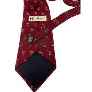 May include: A deep red necktie featuring a repeating pattern of small blue and gold geometric shapes. The tie includes a white label with the text Heritage and a black tag stating Made in Costa Rica of 100% silk imported fabric.