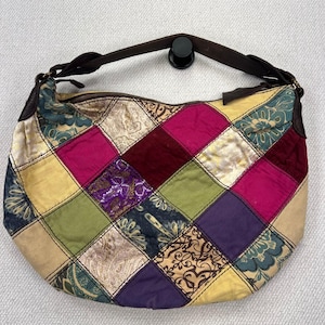 Puede incluir: Un bolso de estilo hobo con un diseño de patchwork de paneles de tela en forma de diamante en tonos magenta, verde oliva, morado, crema y azul verdoso. El bolso incluye una correa de cuero marrón oscuro y un cierre de cremallera superior.