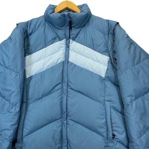 May include: A medium blue puffer jacket featuring a chevron-patterned light blue stripe across the chest. The jacket has a high collar, a full-length front zip closure, and two side pockets. The quilted design provides a textured appearance.