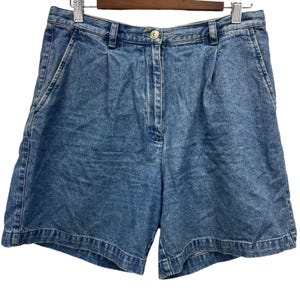 May include: Light blue denim shorts with a button closure and pleats. The shorts have front pockets and a classic, casual style. The garment is hanging on a wooden hanger.