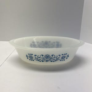 May include: A white, vintage Pyrex bowl with a blue floral pattern. The bowl has a rounded shape with a slightly flared rim and handles. The blue floral design is repeated around the bowl's exterior. This bowl is suitable for serving or food preparation.