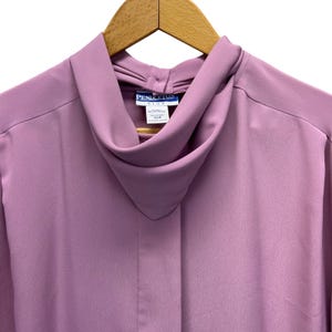 Peut inclure: Un chemisier mauve avec un col roulé, suspendu à un cintre en bois. Le haut à manches longues porte une étiquette indiquant "Pendleton Plus". Le tissu semble lisse et légèrement brillant. Taille 22W.