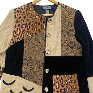 May include: A long-sleeve jacket featuring a patchwork design of black velvet, tan fabric, leopard print, and gold-toned paisley patterns. The jacket has a round neckline, four toggle-style buttons, and a label reading Indigo Moon.