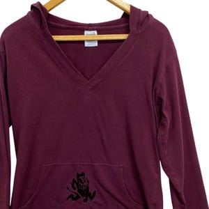 Op de afbeelding: Bordeauxkleurige hoodie met een V-hals en een voorzak. De sweatshirt heeft een zwarte afbeelding van een duivel. De top met lange mouwen hangt aan een houten kleerhanger.