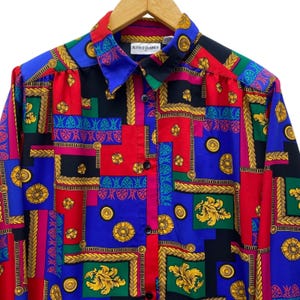 May include: A button-down shirt with a vibrant, geometric pattern in red, blue, black, green, and pink. The shirt features gold-colored floral and rope-like designs. The collar has the brand name "ALFRED DUNNER".