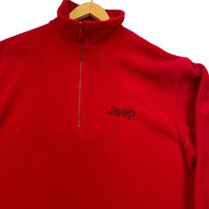 May include: A bright red quarter-zip fleece pullover featuring a high collar and a matching red zipper. The word Jeep is embroidered in black thread on the left chest area. The garment is displayed on a wooden hanger against a white background.