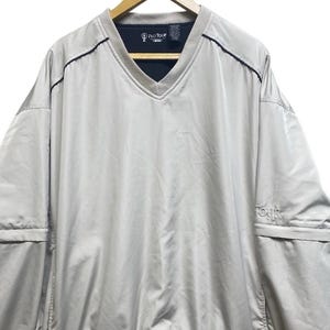 May include: A light gray Pro Tour golf jacket with a V-neck collar and long sleeves. The jacket has a navy blue interior collar and the word "TOUR" is embroidered on the lower right side. The jacket is hanging on a wooden hanger.