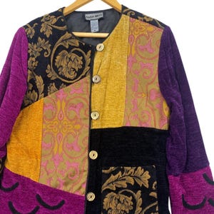 May include: A long-sleeve jacket featuring a patchwork design of purple, mustard yellow, and black fabric panels. The garment includes a black and gold floral brocade pattern, a pink and gold scrollwork print, and five round buttons.
