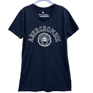 May include: A navy blue t-shirt with a white polka dot pattern. The shirt has a white Abercrombie logo, arched across the top, and a circular emblem below that reads "Eighteen Ninety Two New York".