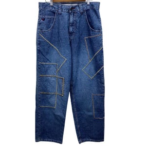 May include: Blue denim jeans with geometric patchwork design. The jeans feature a classic five-pocket design and a button and zipper closure. The patchwork is stitched with yellow thread, adding a contrasting detail to the garment.