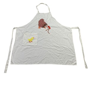 May include: A white apron featuring a brown and white polka-dot hen appliqué with a red comb and beak, pecking at a brown worm. A square pocket on the left side displays a yellow chick appliqué with an orange beak and feet, with neck and waist ties.