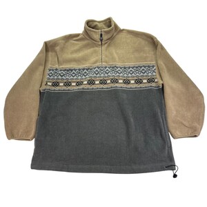 May include: A tan and charcoal gray fleece pullover featuring a quarter-zip mock neck collar. The garment displays a horizontal band across the chest with a geometric pattern in shades of black, white, and light blue. It has long sleeves.