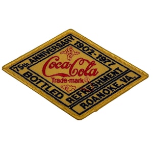 May include: A diamond-shaped embroidered patch with a gold background and black border. It features the red Coca-Cola logo, the text 75th ANNIVERSARY 1902-1977, BOTTLED REFRESHMENT, and ROANOKE, VA. The design includes small red decorative flourishes.