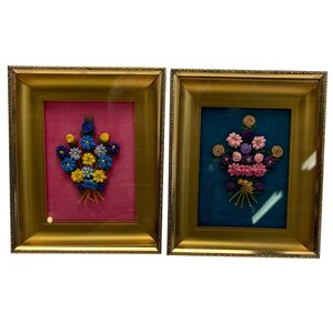 Puede incluir: Dos obras de arte florales enmarcadas. Una presenta un ramo de flores azules, amarillas y blancas sobre un fondo rosa. La otra muestra un ramo de flores rosas y moradas sobre un fondo turquesa. Ambas están en marcos dorados.