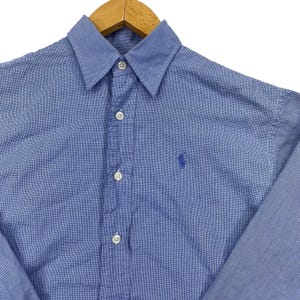 Puede incluir: Una camisa de manga larga con botones en un patrón de microcuadros azul y blanco. La camisa presenta un cuello puntiagudo, una tapeta frontal con botones blancos y un pequeño logotipo bordado en azul de un jugador de polo en el pecho.