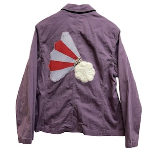 May include: A purple jacket with a decorative patch on the back. The patch features a red and white fan design, a blue and white checkered pattern, and a white flower. The jacket has a black collar.