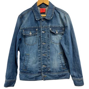 Puede incluir: Chaqueta vaquera azul con cierre de botones, dos bolsillos en el pecho y dos bolsillos laterales. La chaqueta tiene cuello y detalles de costura amarillos. Una etiqueta roja es visible en el cuello.