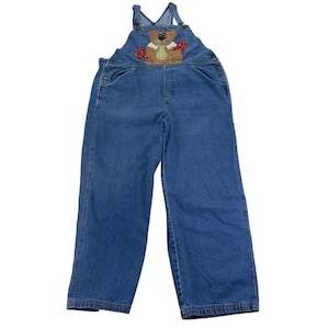 May include: Blue denim overalls featuring a brown teddy bear appliqué on the chest. The bear wears a green and white checkered vest and is surrounded by red star-shaped accents. The garment includes adjustable shoulder straps and side buttons.