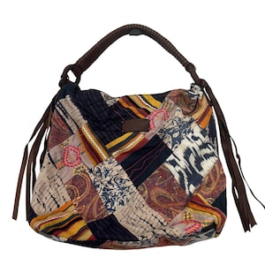 Puede incluir: Un bolso tipo hobo con diseño de patchwork que presenta varios patrones de tela en tonos azul marino, amarillo mostaza, marrón y crema. El bolso incluye un asa trenzada marrón, borlas laterales de cuero marrón y un parche de cuero marrón.