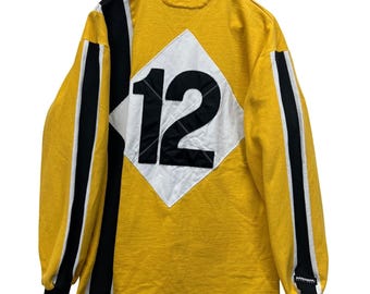 Puede incluir: Camiseta amarilla de manga larga con detalles en negro y blanco. La parte trasera presenta un rombo blanco con el número 12 en negro. Rayas negras recorren las mangas. El cuello es blanco.