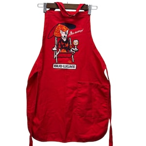 May include: Red apron with a large pocket and tie strings. The apron features a graphic of a dog in a beach chair under an umbrella, holding a glass of beer. The Bud Light logo is printed below the graphic.