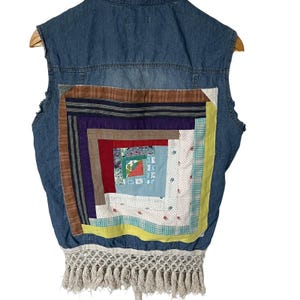 Könnte beinhalten: Eine ärmellose Jeansweste mit einem Patchwork-Quilt-Design auf dem Rücken. Der Quilt zeigt eine Vielzahl von Stoffen in Rot, Blau, Gelb und Weiß. Der untere Teil der Weste hat eine cremefarbene Fransen.