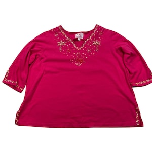 May include: A bright fuchsia three-quarter sleeve top featuring a V-neckline embellished with gold and pink sequins in the shape of palm trees and a flamingo. The sleeve cuffs and side hem slits are also decorated with matching gold and pink sequins.
