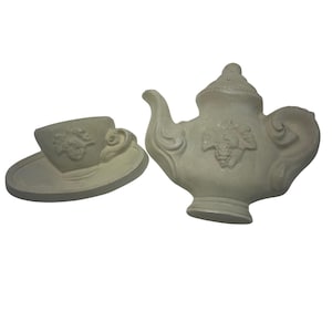 Könnte beinhalten: Ein hellgraues Keramik-Teeservice, bestehend aus einer Teekanne und einer Tasse. Die Teekanne hat einen gebogenen Ausguss und Henkel, mit Deckel und Traubenmuster. Die Tasse steht auf einer Untertasse, ebenfalls mit Traubenmuster. Das Set ist wahrscheinlich dekorativ.