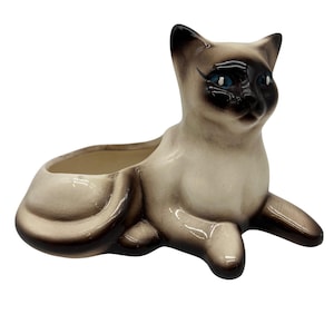 May include: A ceramic Siamese cat planter featuring a cream-colored body with dark brown points on the ears, face, paws, and tail. The cat has bright blue eyes and a glossy finish, with an open basin on its back for holding small items or plants.
