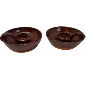 May include: Two brown ceramic candle holders with a handle on the side. The candle holders have a circular base and a small circular center for the candle. The candle holders are glazed with a glossy finish.