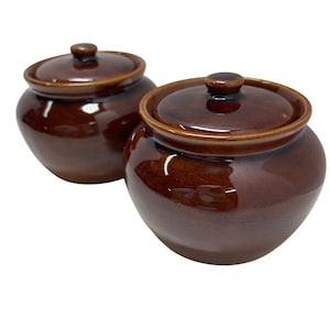 May include: Two brown ceramic jars with lids. The jars have a glossy finish and a rounded shape. The lids have a small knob on top. The jars are likely for food storage or decorative use.