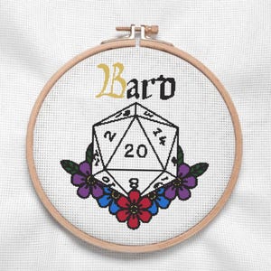 May include: Cross-stitch embroidery in a wooden hoop features the word "Bard" above a white D20 die with the number 20. Purple, red, and blue flowers surround the die, set against a white fabric background.
