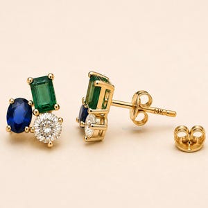 Può includere: Un paio di orecchini a lobo in oro 14K con un gruppo di tre pietre preziose: una pietra verde taglio smeraldo, una pietra blu taglio ovale e una pietra chiara taglio rotondo. Il perno è marchiato 14K e include una chiusura a farfalla in oro abbinata.