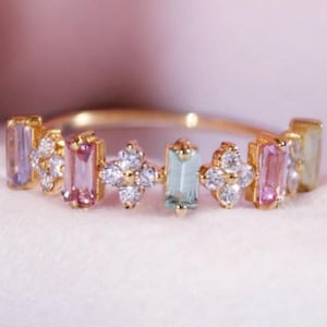 May include: A gold ring featuring alternating rectangular and floral designs. The rectangular designs showcase pastel-colored gemstones in shades of purple, pink, and green. The floral designs are adorned with small, clear gemstones.