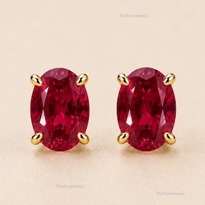 May include: A pair of oval-cut deep red gemstone stud earrings set in yellow gold. Each stone is secured by four prongs, showcasing a faceted surface that reflects light. The earrings are identical in shape and color, featuring a polished gold setting.