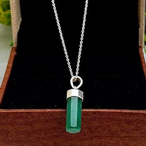Op de afbeelding: Een zilveren ketting met een groene edelsteen hanger. De hanger is een facetgeslepen, smaragdgroene cilinder met zilveren details. De ketting wordt gepresenteerd in een bruine sieradendoos met een zwart interieur.
