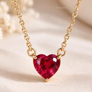 Könnte beinhalten: Eine goldene Halskette mit einem herzförmigen roten Edelsteinanhänger. Der Anhänger ist mit zwei kleinen goldenen Ringen an der Kette befestigt. Der Text TheFunJewels ist in kleiner Schrift oben und unten am Anhänger deutlich zu erkennen.