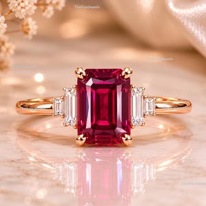 Peut inclure: Une bague en or rose ornée d'une grande pierre précieuse rouge foncé taille émeraude au centre, flanquée de deux petites pierres claires taille baguette de chaque côté. L'anneau est en or rose poli et le texte TheFunJewels est visible.