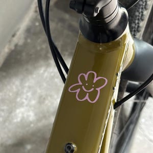 Könnte beinhalten: Nahaufnahme eines olivgrünen Fahrradrahmens mit einem rosa Blumenaufkleber. Der Aufkleber zeigt ein lächelndes Gesicht. Schwarze Kabel und ein schwarzer Vorbau sind ebenfalls sichtbar.